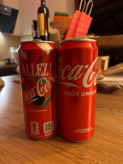 Two red Coca-Cola cans sit stacked on a wooden surface. The top can has the classic Coca-Cola logo and the text "GOÛT ORIGIN". It also has the text "Délicieux & Rafraîchissant". The lower can has a football-themed design, featuring a soccer ball with the Coca-Cola logo. Text on this can reads "ALLEZ!" and "GOÛT ORIGINAL". Near the bottom of the can are symbols, including a QR code and text indicating FIFA partnership. The cans appear to be unopened.