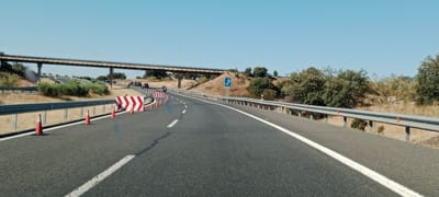The image depicts a highway scene with a clear blue sky. In the foreground, a grey asphalt road with white lane markings curves to the right. Traffic cones and chevron signs are placed along the right side of the road, indicating a lane closure or diversion. A silver guardrail runs parallel to the road. In the middle ground, a concrete overpass spans across the highway. Further back, several cars and a motorcycle are visible on the road, moving away from the viewer. The landscape is characterized by dry, yellowish grass and scattered green trees on a sloping hill. A blue parking sign with a white 'P' is visible on the right side of the road. The scene appears to be on a highway near Jaraicejo, Spain.