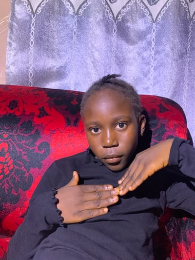 A young African girl, likely a child or pre-teen, is seated on a red and black patterned sofa. She is wearing a black, long-sleeved top with a ruffled neckline. Her hair is dark and pulled back in a small bun. She is looking directly at the camera with a neutral expression. Her left hand is placed over her chest, and her right hand is resting on top of her left. The background consists of a textured gray curtain with a decorative white trim. The lighting is somewhat dim, casting shadows on the girl's face and arms. The setting appears to be indoors, possibly a living room in a home. The provided location information is "Russo, Nigeria."