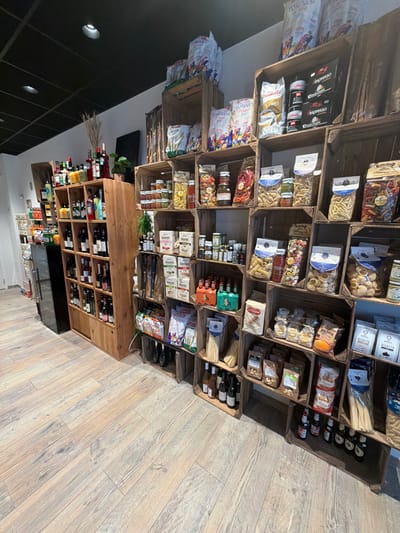 A store display of Italian products features wooden crates stacked with pasta, olive oils, and sauces. A lower cabinet houses wine and other beverages. The scene is bathed in natural light, highlighting the rustic textures of the wooden displays and the colorful packaging of the products. A wooden floor and contrasting dark ceiling complete the store's aesthetic.