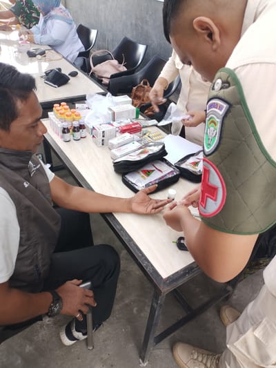 In Cotgadong, Indonesia, a medical service is being provided outdoors. A man, seated and wearing a dark vest over a white shirt, extends his left hand to have his blood glucose tested by a person wearing a light-colored uniform with an emblem and a red cross on their shoulder. The test involves a lancet and a small device. On a table in front of them are various medical supplies, including boxes of medication, bottles of liquid, and test strips. Other individuals, one in a military uniform and another in a white lab coat with a headscarf, are visible in the background at another table. The environment appears to be an open area with a concrete floor and a wall in the background.