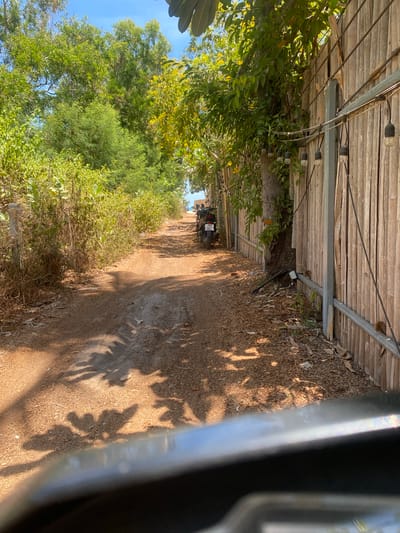 The image captures a sunlit, unpaved dirt path in a rural or semi-rural setting, likely Moo 1, Thailand, given the context and visible license plate. The path extends into the distance, appearing to curve slightly.

On the left side of the path, a tall fence or wall constructed from horizontal wooden planks or bamboo slats runs parallel to the path. Metal support beams reinforce this structure. Along the top of this fence, a string of decorative outdoor lights with clear, exposed bulbs is strung. Green vines and foliage are seen growing on and around the wooden structure, blending with the natural environment.

To the right of the path, a dense expanse of green trees and bushes forms a natural barrier. Some of these trees exhibit yellow blossoms, adding a touch of color. A clear, bright blue sky is visible above the tree canopy.

On the dirt path, in the mid-ground, a black motorcycle is parked or stopped. Its license plate is partially visible, showing Thai script and the numbers "987," confirming the Thai location. The ground itself is covered in dry leaves and dirt, with subtle indentations from tire tracks or footsteps.

Strong, angular sunlight casts prominent, elongated shadows of trees and foliage across the path, suggesting it is either morning or late afternoon. The overall weather appears clear and sunny. A blurred, metallic, possibly curved object occupies the immediate bottom right foreground, indicating the perspective from within a vehicle or behind a barrier. No people are visible in the scene. The atmosphere is peaceful and natural.