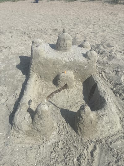 The image shows a sandcastle on a beach. The sandcastle is constructed from damp sand and has a central structure with turrets and a moat-like depression in front. Two feathers are stuck upright in the sand on either side of the central structure, and a small orange object, possibly a cigarette butt, is embedded in the sand on top of the central structure. The sandcastle is surrounded by sand, with some small shells and pebbles visible. The background consists of more sand and a hint of vegetation and a fence in the distance. The lighting suggests it is daytime, with shadows cast by the sandcastle. The location is identified as the city of Rewa, Poland.