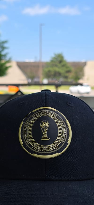 A close-up, eye-level, vertical outdoor shot captures the top of a black baseball cap. The cap features a circular, gold and black emblem of the FIFA World Cup trophy, surrounded by text reading "FIFA WORLD CUP 26TM". The cap is situated in the foreground, with a blurred background of a bright blue sky with white clouds, and a distant, blurred cityscape of buildings and trees. The overall impression is of an outdoor setting, possibly a park or public area, with a focus on the cap's emblem. The exact location in Pickering, Canada is not discernible from the provided image.