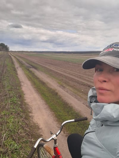 A selfie-style image captures a person on a bicycle in a vast rural landscape, identified as Treshchevka, Russia. The individual, seen from the chest up, wears a light blue or grey hooded jacket and a grey baseball cap. Their face is partially visible beneath the cap, with a neutral expression as they look directly at the camera. A red-outlined text or logo, possibly reading "...ADE RED...", is discernible on the side of the cap.
The person is positioned on a red bicycle, with its silver handlebars and black grips prominent in the lower and central foreground. The setting is dominated by expansive, flat agricultural fields featuring distinct furrows, suggesting tilled or recently planted land. A dirt path or track stretches into the distance, delineating the fields. Sparse green vegetation lines the path and is scattered within the fields. A faint line of trees marks the far horizon. The sky is overcast with varying shades of grey, indicating a cloudy daytime. The scene conveys a sense of serene travel through open countryside.