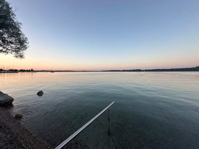 This image depicts a serene lake scene at dusk, likely on the shore of Lake Chiemsee in Prien am Chiemsee, Germany. The sky transitions from a soft gradient of pale blue at the top to warm hues of pink and orange near the horizon, indicating the sun has recently set. A lone tree branch with sparse leaves is visible on the left side of the frame, casting a slight shadow.
The water itself is calm, with subtle ripples reflecting the fading light. Several rocks are present in the shallow water near the shore, some partially submerged. The shoreline is composed of pebbles and small stones. In the foreground, a metallic railing, appearing to be part of a dock or entry point into the water, is angled from the bottom right corner towards the lake.
In the distance, the far shore of the lake is lined with a dense row of trees. A small cluster of sailboats and masts can be seen on the left side of the horizon, suggesting a marina or harbor. One or two small boats are also visible further out on the water. The overall atmosphere is tranquil and peaceful, capturing the quiet beauty of the lake at twilight.