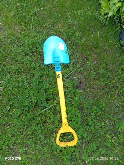 In Wilczyce, Poland, a blue and yellow toy shovel lies on a patch of green grass dotted with small clover leaves and some scattered weeds. The shovel is positioned diagonally across the frame, with the blue scoop at the top and the yellow handle extending downwards towards the bottom right corner. The grass appears slightly uneven and some dry patches are visible. A few small purple and yellow wildflowers are scattered in the grass. A white label is affixed to the blue part of the shovel. The bottom left corner of the image shows a watermark reading "POCO C75" and the bottom right corner displays a timestamp "09.06.2026 10:52". No people or animals are present in the scene.