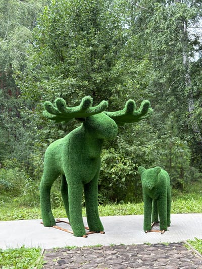 This image depicts two topiaries, one resembling a large moose and the other a smaller bear, situated outdoors on a paved pathway in the city of Zarechnyy, Russia. The topiaries are constructed from green, artificial turf and are positioned in a forest-like environment with abundant trees and foliage in the background. The larger moose topiary features prominent antlers, while the bear topiary is smaller and more compact. The pathway appears to be made of light-colored concrete slabs, bordered by a decorative arrangement of circular wood slices. The scene is set during daylight, with natural light illuminating the subjects and their surroundings.