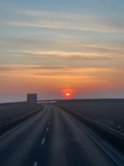 The image depicts a two-lane highway at sunrise or sunset, presenting a view from a moving vehicle. The scene is located near the city of Slaný, Czechia. The sky dominates the upper half, transitioning from light blue at the zenith to vibrant orange and deep red near the horizon. A prominent, intensely red, partially obscured sun is positioned low on the horizon, slightly right of the road's center line.
The asphalt road curves gently to the left in the foreground, then turns distinctly to the right in the mid-ground, receding into the hazy distance towards the sun. White dashed lane markers are visible on the road surface. Guardrails line both sides of the road. On the right, a small white and red reflective roadside marker is partially visible in the lower corner. On the left, further down the road, a large, rectangular, dark grey sign or barrier stands adjacent to the roadway. A bridge or overpass structure spans horizontally across the road in the mid-distance, positioned beneath the horizon and to the left of the sun. The surrounding environment appears as dark, low-lying land, possibly fields, obscured by atmospheric haze or low-lying fog, particularly towards the horizon. No people or active vehicles other than the presumed source of the image are visible.
