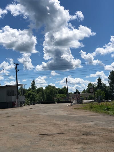 The image depicts a railway crossing in Izdeshkovo, Russia, on a clear day with a vibrant blue sky dotted with fluffy white and gray clouds. In the foreground, a rough, asphalt road shows signs of wear with numerous potholes. To the left, a small, white building with a covered porch and visible stairs is situated near a utility pole carrying multiple overhead wires. A red and white striped barrier is visible at the railway crossing. Further into the scene, a sign displays "4.5 M" within a red circle, indicating a height restriction. Dense green trees line the background, bordering the railway tracks. The overall scene is a functional, rural railway crossing.