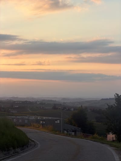 The image shows a vertical view of the Castiglione Tinella, Italy, landscape. In the foreground, a winding asphalt road is bordered by grassy vegetation. Adjacent to the road, there is a multi-story building with visible windows. The distance reveals a hazy landscape with houses scattered among the terrain. In the sky, elongated clouds are tinted with hues of orange and grey, suggesting dawn or dusk.
