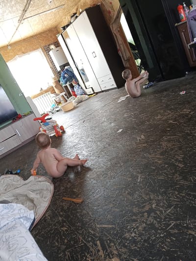 The image depicts an indoor domestic scene in Teykovo, Russia, featuring two young children within a functionally furnished and somewhat cluttered room. In the mid-right foreground, an older child, appearing to be a boy with short, light-colored hair, is seated directly on the dark floor. He is unclothed, facing away from the camera, with his legs extended forward and his hands holding his feet in a seated yoga-like pose. Adjacent to him on the floor are a small blue toy car, a crumpled piece of white material (possibly paper or fabric), and a small red and white toy. In the left foreground, a younger child, identified as a baby or toddler with light-colored hair, is also seated directly on the dark floor, unclothed, and facing away from the camera. The child leans slightly to the right and holds a small, orange, cylindrical object in their right hand. Near this child, a cream-colored blanket with a blue and white "Star Wars" pattern is partially visible. A wooden spoon lies on the floor to the child's right. The floor throughout the room is constructed from dark-colored Oriented Strand Board (OSB) or a similar particle board, exhibiting a textured, splintered appearance. The walls and ceiling are predominantly light-colored OSB, giving the room an unfinished or utilitarian aesthetic. Exposed electrical wiring runs along the ceiling, where a single fluorescent light fixture is mounted. On the left side of the room, a large window provides significant natural light, partially obscuring the view outside with what appears to be a sheer white curtain or plastic sheeting. A white radiator is visible beneath the window. Below the window and to the left, a low, light-colored wooden media console or cabinet with drawers supports a dark-screened electronic device, likely a television. Further into the room, on the right side, a large white wardrobe with mirrored doors and multiple drawers dominates the space. A pile of laundry or clothes is visible on a surface next to this wardrobe. To the right of the wardrobe, a dark, reflective panel, possibly part of a refrigerator or another large cabinet, is present. A smaller, darker wooden cabinet sits to the right of this panel, with an orange plastic bottle (resembling a cleaning product container) and a purple towel on its surface. Adjacent to the window wall, a white wall-mounted boiler or water heater is installed over a section of wall adorned with