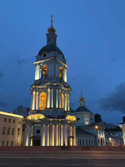 The image showcases a tall, ornate church tower against a twilight sky. The tower has a distinctive blue and white facade, with multiple levels featuring columns and arched openings illuminated by warm light. A green dome sits atop the tower, capped with a golden spire and a cross. To the right of the main tower, a smaller, similarly styled structure is visible. In the background, city buildings line the street, illuminated by streetlights. The scene is set in Перовское сельское поселение, Ukraine.