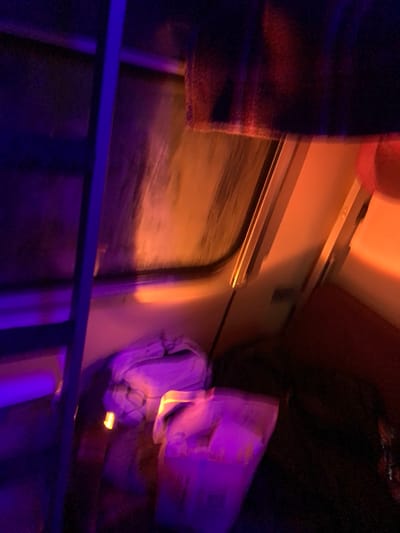 A blurry shot inside what seems to be a train compartment. The window shows an indistinct landscape in motion. A curtain hangs on the left, next to the window. The interior lighting is a mix of orange and purple hues, obscuring details. A luggage rack and overhead storage are partially visible. A blue railing is in the foreground. The overall image is dark and difficult to discern.