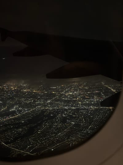 This is an aerial nighttime view of a city, seen through an airplane window. The city lights are a dense network of yellow and white dots, indicating a sprawling urban area. The lights are particularly concentrated in some areas, suggesting downtown districts or densely populated neighborhoods. Some roads or highways can be faintly discerned by the lines of lights that run along them. The scene is partially obscured by the silhouette of an airplane wing in the foreground. The atmosphere appears clear enough to see the city lights distinctly, though there may be a slight haze or clouds visible in the distance, catching some of the city's glow. There are no visible people, specific landmarks, or discernible actions. The overall impression is one of a large, active city at night from an elevated perspective.