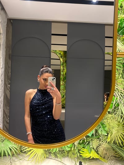 A woman is taking a selfie in a mirror. She is wearing a dark blue, sequined dress and jewelry on her wrists, ears, and fingers. The mirror has a gold frame, and behind the mirror is a wall decorated with lush green plants and gray paneling. The reflection also shows a white panel. Based on the architectural style and decor, the location appears to be an upscale interior, possibly a restaurant or event venue. However, there is no information to place this in the city of Masis, Armenia.