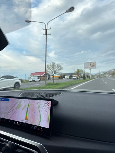 The view from inside a car shows a road leading towards Yerevan, Armenia, with a GPS navigation display indicating the driver is on the Yerevan-Abovyan Highway and approximately 28 km from the city center. The surrounding environment includes roadside infrastructure such as streetlights and billboards. A white car is visible in the foreground. The sky is cloudy. The GPS display shows a speed limit of 90 and the current speed as 82.