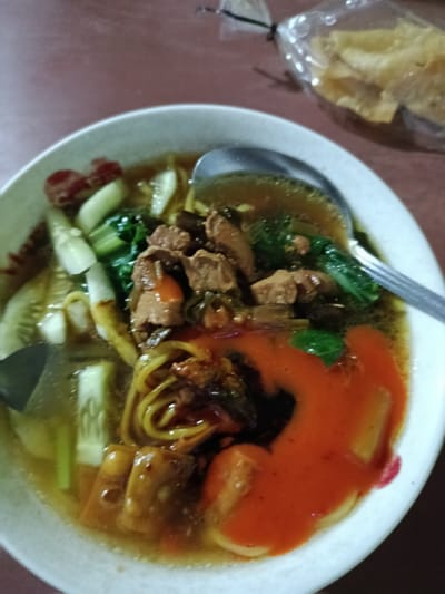 A close-up view depicts a white ceramic bowl containing a noodle dish, resting on a dark brown table surface in Arjasa, Indonesia. The bowl is filled with yellow noodles in a light brown broth, accompanied by chunks of dark brown meat (likely chicken), green leafy vegetables, and sliced cucumbers. A significant portion of bright orange-red chili sauce or sambal is drizzled over the noodles on the right side of the bowl. A silver-colored spoon is partially submerged in the broth on the upper right. In the upper right background, a clear plastic bag containing light-colored, wavy crisps or crackers is visible, tied at the top. No people are present, and no active interactions are depicted.