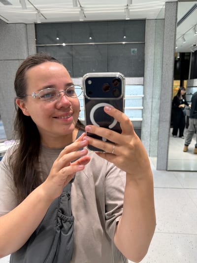 A young woman with dark brown hair pulled back and wearing eyeglasses with a decorative embellishment on the bridge is taking a selfie in a store. She is wearing a light brown t-shirt and a gray crossbody bag. She is holding a smartphone in her right hand, which is angled to capture her reflection in a large mirror. Her left hand is near her face, and she is wearing a thin silver ring on her left index finger. In the background, two other people are visible in the mirror, one wearing a black top and the other wearing a black jacket and gray pants. The store appears to be a retail environment with shelves and lighting fixtures visible. The location is a store in Chuo, Japan.