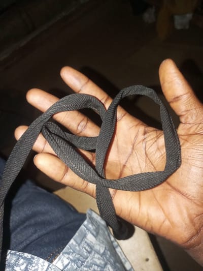 A close-up, slightly overexposed shot shows a person's open palm in Agbara, Nigeria, holding a black fabric cord looped into a heart shape. The cord has a ribbed texture. The background is dark and out of focus, with hints of wooden furniture and possibly a yellow object. The person's hand is dark-skinned with visible lines and creases. The person is wearing dark denim jeans and a patterned fabric, possibly a shirt or accessory, partially visible at the bottom of the frame. The lighting is dim, suggesting an indoor setting at night.