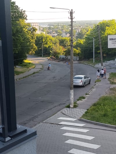 The image shows a street scene in Fastiv, Ukraine. In the foreground, a dark metal pillar is visible on the left. A pedestrian crossing with white stripes is laid on a gray brick sidewalk in the lower part of the image. The street is asphalt, with visible cracks and patches. A silver sedan is parked on the right side of the street. Three people are walking on the sidewalk to the right of the car. Further down the street, a person is pushing a stroller. Trees line the sides of the road, and in the background, there are houses and buildings, as well as utility poles and overhead wires. The sky is bright and overcast. A billboard with text in Ukrainian is visible on the right side of the image, behind the utility pole.