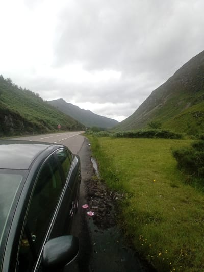 A dark colored sedan is parked on the side of a two-lane road. The road curves to the right and leads into a valley between two green, mountainous hills. The sky is overcast and grey, with thick clouds covering the sun. The hills are covered in lush green grass and dotted with trees. A small patch of yellow flowers is visible in the grass on the right side of the road. Two pink flip-flops are lying on the wet ground next to the car. The scene appears to be in a remote, natural area. The image is taken from a slightly elevated perspective, looking down at the car and the road.