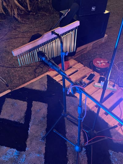 The image depicts an outdoor musical performance setup at night in Murera ward, Kenya. The foreground features a black and light grey patterned area rug laid on dry earth. Two black tripod microphone stands are positioned on the rug. One stand holds a black dynamic microphone. Behind this, a wooden bar chimes instrument, comprising multiple vertical metallic tubes, is mounted on a stand.

To the right, on two stacked wooden pallets, rests a large, black EV (Electro-Voice) speaker box. Adjacent to the speaker, a red cable reel and a white power strip with multiple outlets are visible. Two smartphones are placed on the pallets, one connected to the power strip.

In the upper left background, an obscured portion of an electric guitar neck is visible behind the chimes. Indistinct, dry vegetation is present in the far background. The scene is illuminated by artificial light, casting a blue and purple ambient hue over the equipment and ground. No individuals are present in the frame.