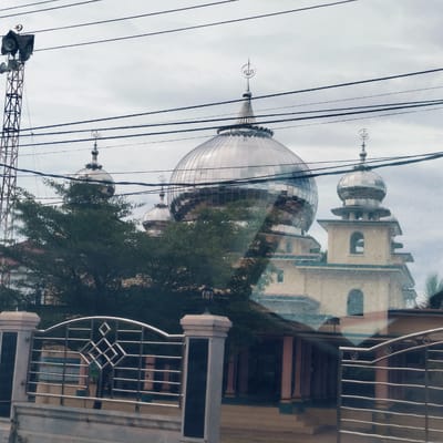 The image captures a modern mosque under an overcast sky, seen through what appears to be a window or glass, evidenced by reflections. The central focus is a large, shimmering silver or chrome-domed mosque, flanked by smaller domes and minarets of similar metallic material. The mosque building itself has light-colored walls, possibly yellow or beige, with arched windows and blue trim.

To the left of the main mosque structure, a tall, metal lattice tower stands, equipped with multiple loudspeakers and topped with a crescent moon and star symbol, typically associated with Islam. Numerous black utility cables or power lines traverse the upper portion of the frame horizontally, creating visual lines across the sky and mosque domes. Green trees and foliage are scattered throughout the midground, partially obscuring parts of the mosque and its surroundings.

In the foreground, a decorative metal fence or gate, likely made of stainless steel, features geometric patterns and is connected to a stone pillar. Behind this fence, another building with reddish-pink pillars is partially visible. Reflections on the large mosque dome show fragmented views of the sky and potentially other elements of the environment.

Notable details include the Arabic calligraphy "Allah" (الله) visible as a finial on top of the largest dome and some smaller domes, and the crescent moon and star on the loudspeaker tower. Through the fence, a sign with the partial text "PANITIA QURBAN" (Qurban Committee) can be discerned. The scene is static, depicting a daytime setting with cloudy weather, characteristic of Barat Yaman, Indonesia.
