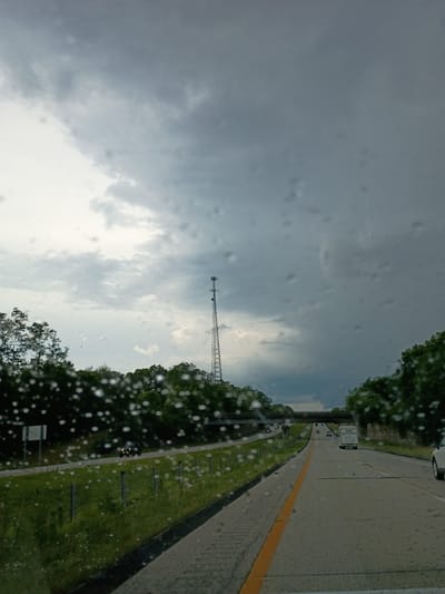The image depicts a view from inside a vehicle, looking out at a highway scene on a cloudy day. The perspective is from the passenger side of the car, with raindrops visible on the windshield, suggesting it might be raining or has recently rained. The highway stretches into the distance, with one lane clearly marked by a yellow line. Several vehicles are visible on the road, including cars and a truck.

To the left of the highway, there is a grassy verge bordered by trees. Further in the distance, a tall communication tower stands against the sky, which is filled with dark, heavy clouds, indicating a potentially stormy or overcast weather condition. The time of day is not clearly discernible, but the general lighting suggests it could be afternoon.

On the right side of the highway, the road surface is visible, and it appears to be wet. The overall scene conveys a sense of travel and perhaps a slightly somber mood due to the cloudy weather. There is a white truck with text on its side that is partially legible, reading "LAMAR" followed by a phone number. Another sign is also visible near the highway. The location is cued as Frankfort, United States.