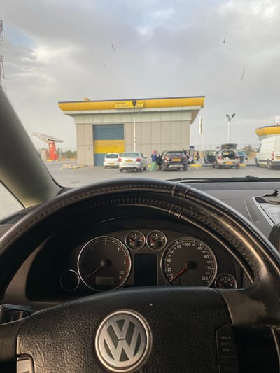The image shows a view from inside a car, looking through the windshield. In the foreground, the steering wheel of a Volkswagen is visible. Beyond the steering wheel, a building with a yellow awning and a blue and yellow striped garage door is situated. Several cars are parked in front of the building, and some people are interacting with them. The scene appears to be a service station or a car wash. The license plates on the cars are partially visible, suggesting a location in Algeria. The sky is overcast. The time on the car's dashboard clock reads 2:16. The odometer shows 360386. The location is identified as Mocta Douz, Algeria.