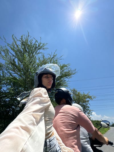 The image depicts two individuals riding on a motorcycle under a bright, clear blue sky with the sun high and shining intensely. The person in the foreground, on the left, is a woman wearing a black helmet and a light-colored, sheer long-sleeved top over a ribbed tank top, and distressed jeans. She is looking towards the camera and sticking her tongue out. A patterned scarf is visible around her neck. Behind her, another person, likely male, is wearing a black helmet and a pink t-shirt. A third person, only partially visible, is wearing a gray t-shirt and a white helmet. The background consists of lush green trees, power lines stretched across poles, and a paved road curving to the right. The overall setting suggests a rural or suburban area, potentially on the outskirts of Hong Son, Vietnam, given the vegetation and the presence of power lines. The perspective is from the motorcycle, looking forward and slightly to the side.