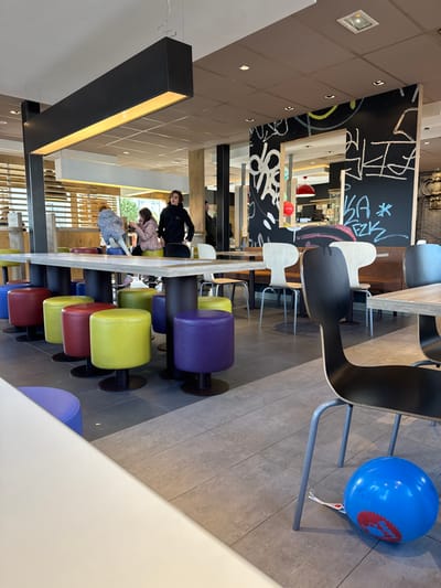 This image depicts the interior of a fast-food restaurant, identified as a McDonald's, located in Oberhausbergen, France. The scene is observed from a slightly elevated perspective, looking towards the mid-ground.
**Environment:**
The dining area features a light grey tiled floor. The ceiling is paneled in a light brown or beige hue, with recessed lighting fixtures. A prominent, elongated black rectangular light fixture with an illuminated underside hangs from the ceiling in the mid-left. Dark pillars are visible, some adorned with white graffiti-style markings and text, including a section with a mirror-like cutout. Blinds or slatted structures are visible through a large window in the far background on the left. Red hanging dome lights are visible in the distant background.
**Seating and Tables:**
In the foreground, the edge of a white table is visible on the left. In the mid-ground, a long, light-colored rectangular communal table with a dark base is surrounded by a row of colorful, round upholstered stools in blue, red, yellow, and purple, each with a dark circular base. Beyond this, various other tables and chairs are arranged throughout the dining area, including light wood-toned chairs with dark backs and brown upholstered booth seating. In the immediate right foreground, the back and metal legs of a dark chair are prominent.
**People:**
Three individuals are situated around the communal table in the mid-ground. On the far left, a young child with light-colored hair, wearing a patterned jacket, is kneeling on a blue stool. Next to the child, a person with light brown hair, wearing a light-colored top, is seated on a yellow stool, looking down at a device in their hands. To their right, a person with dark, curly hair, wearing a black hooded top, stands beside the table, facing towards the left. A black bag rests on the floor next to this individual. Partially obscured figures are visible further in the background.
**Objects and Actions:**
The child on the stool is positioned as if interacting with the table or the person next to them. The seated person is engaged with a personal electronic device. The standing person appears to be observing the area or waiting. On the floor in the bottom right corner, next to the chair, lies a deflated or partially deflated blue balloon with red text, partially visible as "Happy," indicating a "Happy Meal" balloon. A white ribbon is attached to the balloon.