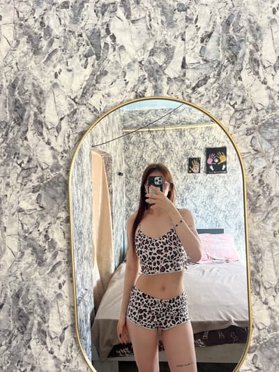 A person is taking a selfie in a bedroom. The person is wearing a leopard print pajama set. They are standing in front of a large, oval mirror with a gold frame. The mirror reflects the bedroom, which includes a bed with pink and beige bedding, a draped curtain, and two framed pictures on the wall. The walls in the bedroom are covered in a marble-patterned wallpaper. The person is holding a smartphone in their right hand, taking the photo. The scene is indoors and appears to be in a domestic setting. It is not possible to determine the location in the city of Blizhnii Khutor, Moldova from the image alone.