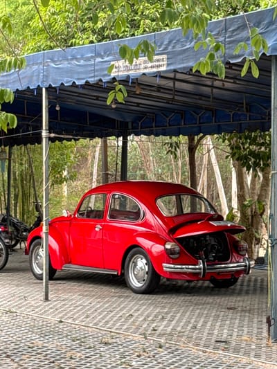 A red Volkswagen Beetle, with its hood open, is parked under a blue tarp structure. A motorcycle is parked behind the car. The location is likely Muang Pattaya, Thailand, due to the presence of Thai script on a sign attached to the tarp structure. The ground is paved with a metal grate material. The scene includes elements of vegetation, with trees and plants visible in the background and foreground.