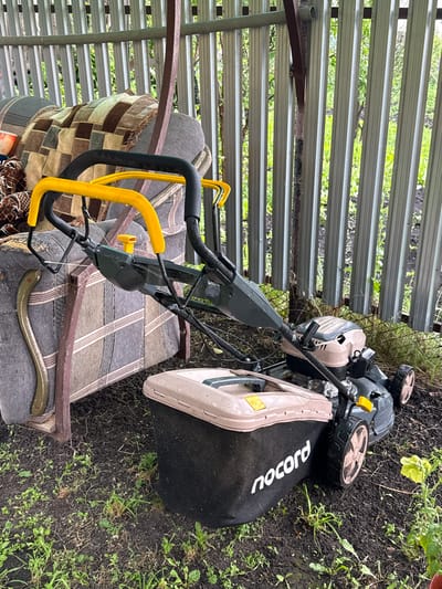In the city of Kandry, Russia, a lawnmower is parked on the ground next to an upholstered seat covered with a checkered blanket. The lawnmower is dark gray with a tan top and a black grass bag that says "nocord" in white. The lawnmower has two yellow handles and black wheels. The upholstered seat is made of striped fabric in brown, grey, and white colors. Behind the lawnmower and seat, a corrugated metal fence is visible. The ground is covered with dark soil and scattered green weeds. The scene appears to be outdoors under a covered area, possibly a carport or shed.