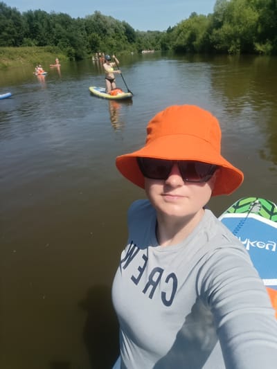 In this medium shot, the photo is taken from a first-person perspective on a paddleboard, looking out over a river surrounded by lush green trees on both banks under a bright blue sky. In the foreground, a person wearing an orange bucket hat and sunglasses is centered, their upper body visible. They are wearing a light gray t-shirt with dark lettering on the front that reads "CREW." Next to them, to the right and partially visible, is a blue and green paddleboard. In the background, several other people are paddleboarding on the river. One woman in a bikini is standing on a paddleboard and paddling. Another paddleboarder is seated. The water is a murky brown, reflecting the trees and sky. The overall impression is of a recreational day on the water. The location is identified as Berkuty, Russia.