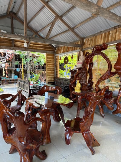 The image shows an outdoor covered seating area with rustic wooden furniture carved from tree roots. The furniture includes a large, irregularly shaped table and several chairs with unique, organic designs. In the background, there are more wooden structures, greenery, and various decorative items hanging from the ceiling and structures, suggesting it may be a shop or a workshop specializing in wooden crafts. The overall impression is of a natural and artistic environment. The location is specified as Hòa Đa, Vietnam.