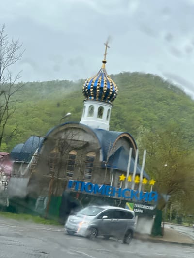 The image shows a building with a blue and gold dome, likely a church, with a cross on top. The structure is under construction with scaffolding visible. A sign in Russian, "ТЮМЕНСКИЙ РАЙОН" (Tyumenskiy District), with yellow stars above, is visible next to the building. A silver car is parked nearby. The background features a green hillside or mountain and an overcast sky. The photo was likely taken from inside a vehicle as evidenced by water droplets on the glass. The location is Tyumenskiy District, Russia.