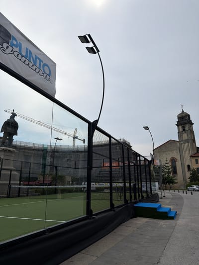 The image shows a paddle tennis court situated in an urban environment in Pisa, Italy. The court itself is green with white boundary lines and a net. It is enclosed by a metal fence. In the foreground, a banner with "PUNTO GOMMA SNC" written on it is visible, along with an image of a tire. This suggests the area might be related to a tire shop or automotive service.

Behind the court, a church with a distinctive bell tower stands prominently, a common architectural feature in Italian cities. The sky is overcast, indicating a cloudy day, and the lighting suggests it is daytime, possibly late afternoon or early morning due to the soft light. There is a construction crane visible in the background, hinting at ongoing development or renovation in the area. A statue of a person is also reflected in the glass walls of the court. The overall scene depicts an outdoor sporting facility within a historically rich urban setting, with elements of modern commercial advertising and ongoing construction.