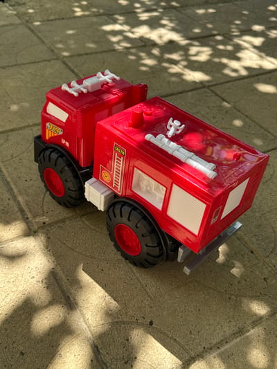 A red toy fire truck is situated on a paved, light-brown tiled surface. The truck has black tires with red rims and features white accents, including what appear to be hoses on top and ladders on the side. The truck has the number "719" and "LP-19" on its front section. The word "CALL 719" is visible on the side of the rear compartment. The scene is illuminated by sunlight, casting dappled shadows from foliage onto the ground and the truck. The setting is an outdoor area.