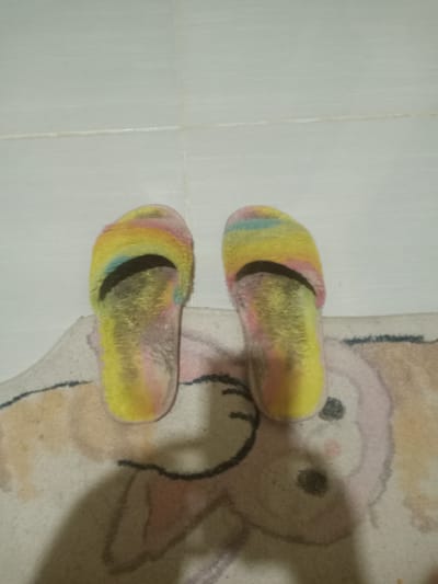 A pair of fluffy, rainbow-colored slippers are positioned on a rug with a cartoon character design, possibly a rabbit. The slippers are worn and appear to have some dirt or dust on them. They are placed on a tiled floor. The lighting suggests an indoor setting. The location is described as Uleeceubrek, Indonesia, though there is no visual information in the image to confirm this specific geographic detail.