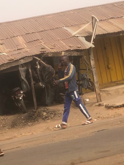 The scene is set in Tukur Tukur, Nigeria, depicting a street adjacent to a rudimentary workshop under a hazy sky. A dark-skinned, bald male, dressed in a blue jacket with yellow sleeve stripes, blue trousers with white side stripes, and two-toned sandals, walks left-to-right. He holds a black smartphone to his right ear, speaking, while his left hand is lowered, possibly holding a small white item. He traverses a dirt shoulder next to a paved road. The background features a single-story structure with a corrugated, weathered brown metal roof, partially supported by wood and weighted with rocks. A white, curved fragment resembling a satellite dish is affixed to the roof on the right. The open shop front reveals a cluttered interior: a dark motorcycle on the left, interwoven metal components or bicycle frames centrally, and a large tire or similar object in the rear. The ground immediately before the workshop is dirt and rubble. A yellow-painted wall is visible to the right of the shop opening. A single foot in a sandal is partially visible on the far left foreground.