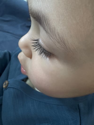 A close-up, high-angle shot depicts the right side profile of a sleeping or resting infant or toddler. The child's skin is fair, with a smooth texture. The right eye is closed, featuring notably long, dark eyelashes. A fine, dark eyebrow is visible above the eyelid. The bridge of a small nose is present. The mouth is slightly agape, revealing a portion of the moist, pink lower lip and tongue. Fine, dark hair is visible on the scalp. The child wears a dark blue garment with a subtle ribbed texture, secured by a visible round, brown button on the upper left side of the frame. The background is an out-of-focus dark blue, consistent with the child's clothing, suggesting a fabric surface. The location in the city of Apshak, Uzbekistan, cannot be determined from the visual information provided within the image.