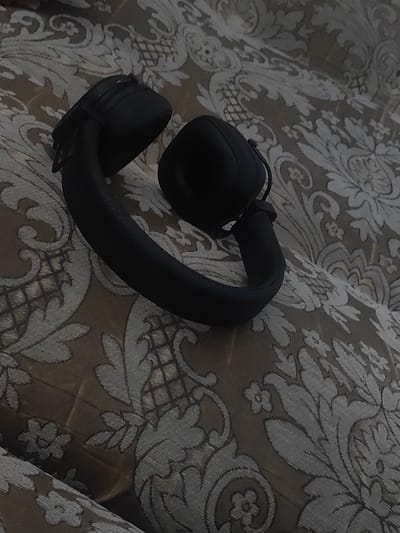 A pair of black over-ear headphones rests on a patterned surface, likely a couch or bed, with a detailed damask-like design in muted tones of brown and grey. The headphones are positioned with the headband curved, suggesting they are laid down. One ear cup is visible, rounded and black, with a slight sheen. The other ear cup is mostly obscured. The background fabric exhibits a repeating floral and scrolling vine motif. The lighting is dim, casting soft shadows and emphasizing the textures of both the headphones and the fabric. The scene appears to be indoors. There are no people or any other objects present in the image. The location in Zhanadaur, Kazakhstan is not discernible from the image itself.
