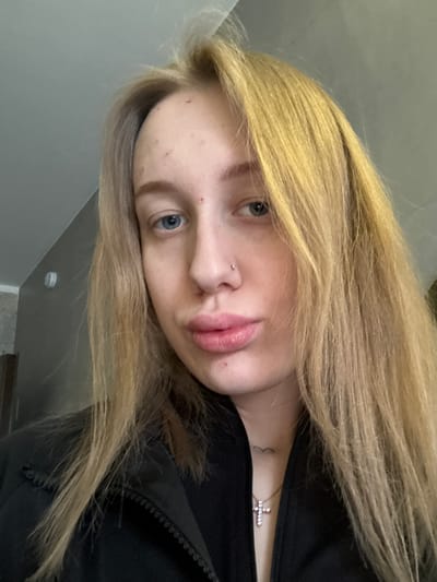 A young woman with long, straight blonde hair and blue eyes is depicted in a close-up portrait. Her head is tilted, and she looks directly at the viewer with a neutral, slightly pensive expression. She has full, pink lips and a small stud piercing on her nose. Her skin shows some visible blemishes.
She is wearing a dark, likely black, jacket with a zippered front, part of which is visible on the right side of the image. Around her neck, a delicate necklace with a small, sparkling cross pendant is worn. A faint, small tattoo, possibly a heart or infinity symbol, is visible on her upper chest, near her collarbone.
The background is a plain, light grey wall, indicating an indoor setting. In the lower right corner of the original image, a white circular object, resembling a smoke detector or a simple light fixture, is visible on the wall. The lighting appears even, suggesting an artificial light source or soft ambient daylight from indoors. There is no visible text in the image.