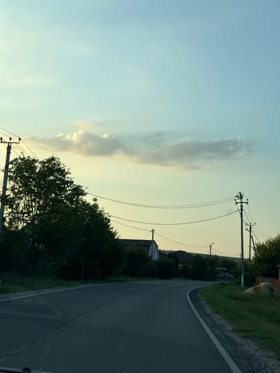 The media file depicts a road in Nizhneatamanskoe, Russia, during what appears to be the late afternoon or early evening. The sky is a pale blue, transitioning to a soft yellow hue near the horizon, indicating the sun is low in the sky. Wispy clouds are scattered across the sky, some catching the golden light.
The road itself is paved and curves gently to the right. It is bordered by a grassy verge on the left and a slightly elevated, rocky embankment on the right. Several utility poles with wires stretch along the roadside, suggesting an inhabited area.
On the left side of the road, there are trees and the roofs of houses visible, indicating a rural or semi-rural setting. The foliage of the trees is dense and green. On the right side, the embankment is covered in grass and scattered rocks, with more trees and vegetation further up the slope. The overall atmosphere is peaceful and tranquil, bathed in the soft light of the setting sun. There is no visible text in the image.