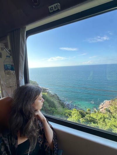 A medium shot depicts a woman inside a moving vehicle looking out a window. The vehicle is likely a train or a bus, based on the interior paneling and window size. The window provides a scenic view of the ocean and a rocky coastline covered in green vegetation. The sky is blue with a few clouds. The woman has long, dark hair and is wearing a patterned shirt. A water bottle sits near the window. A sticker with the numbers "42 41" is visible on the vehicle’s interior. While the image features an ocean view, there is no immediate indication that this scene is in Huế, Vietnam. Further visual or contextual information would be needed to confirm the location.