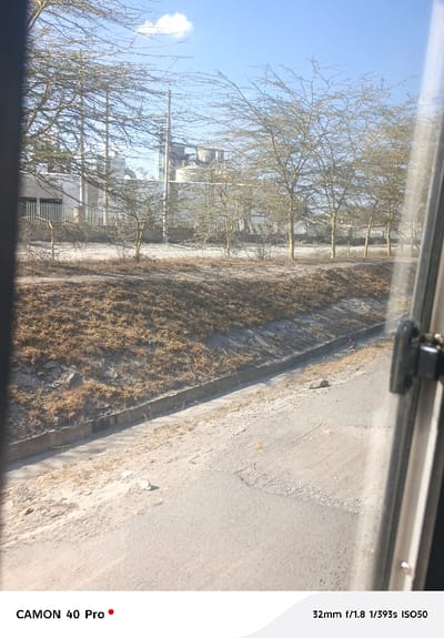 A slightly blurred, sunlit view from a vehicle window shows a flat, sandy landscape. Bare trees line the horizon, obscuring an industrial building with tanks. A dry, grassy bank slopes to a concrete drainage ditch next to a cracked, dusty road. The camera's brand and technical settings are visible at the bottom of the frame. The sky is blue with a single white cloud.