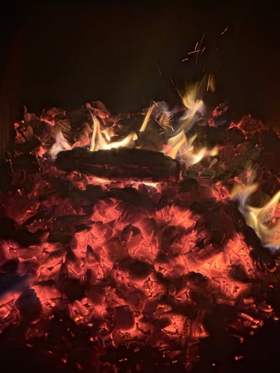 The image depicts a close-up view of glowing embers and burning wood. The scene is dark, with the primary light source coming from the intense orange and red glow of the fire. Several pieces of wood are visible, some partially burned and others engulfed in flames, casting dynamic yellow and white highlights. Small sparks are scattered throughout the upper portion of the image, indicating active combustion. The texture of the embers appears rough and uneven, with the flames creating a sense of movement and heat. The background is a deep black, suggesting a lack of ambient light and focusing all attention on the fire. There is no indication of any specific location within the city of Таганрогский, Russia, or any people present in the image.