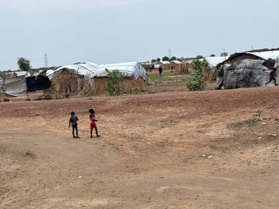 Two young children, one wearing a red shirt and black shorts and the other wearing a red shirt and black pants, walk across a dirt path in what appears to be a refugee camp. Behind them, a collection of makeshift shelters constructed from straw and tarpaulins are visible, suggesting a rural or semi-rural setting. In the background, power lines and sparse trees indicate the presence of infrastructure. The overall scene suggests a developing or impoverished area, likely within or near Ad-Damazin, Sudan, given the context.
